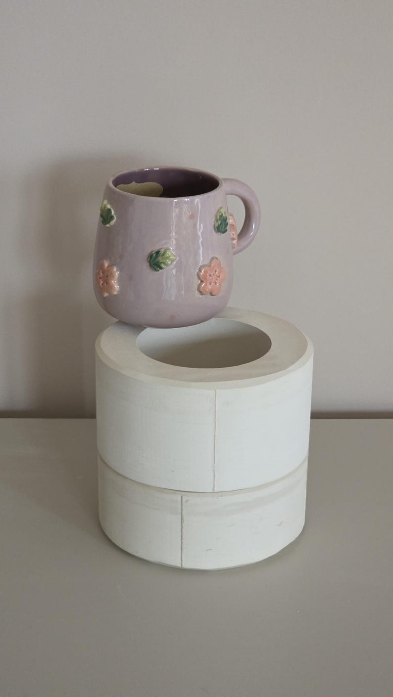 May include: A lavender ceramic mug with a handle, decorated with raised pink and green floral designs. The mug is placed on a white, cylindrical, two-tiered stand. The background is a neutral grey.