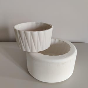 以下が含まれることがあります： 白いセラミック製のプランターが2つ、重ねられています。上のプランターは、テクスチャのあるリブ模様になっています。下のプランターは滑らかです。
