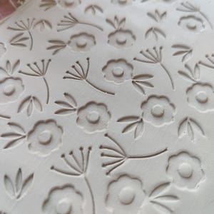 Könnte beinhalten: Nahaufnahme einer weißen Tonfläche mit einem sich wiederholenden Muster aus kleinen Blumen und Blättern. Das Muster entsteht durch das Drücken eines Stempels in den Ton.