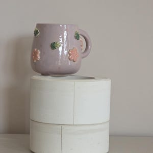 May include: A lilac ceramic mug adorned with pink and green floral details. The mug features a handle and rests on a white, cylindrical, two-tiered stand. The mug is a decorative item.