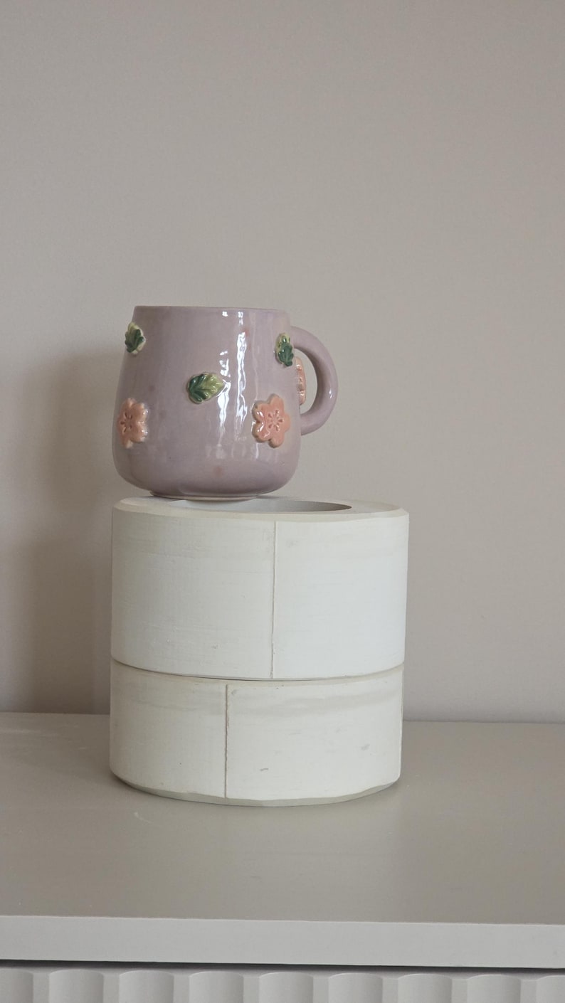 May include: A lilac ceramic mug adorned with pink and green floral details. The mug features a handle and rests on a white, cylindrical, two-tiered stand. The mug is a decorative item.