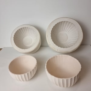 Op de afbeelding: Vier witte keramische kommen, twee zijn mallen en twee zijn afgewerkte kommen. De mallen hebben een geribbeld ontwerp aan de binnenkant. De afgewerkte kommen hebben een geribbeld ontwerp aan de buitenkant.