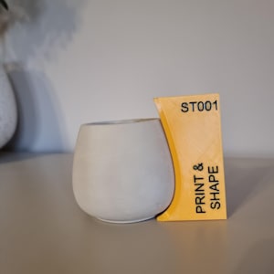 Op de afbeelding: Een gele plastic pot met de tekst "PRINT & SHAPE" en "ST001" aan de zijkant. De pot bevat een witte keramische pot.