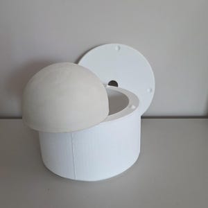 Op de afbeelding: Een witte cilindrische mal met een gladde, matte afwerking en een ronde opening. Een lichtgrijs, koepelvormig object rust gedeeltelijk in de bovenkant van de mal. Een apart, plat, rond wit deksel met een klein gat staat erachter geplaatst.