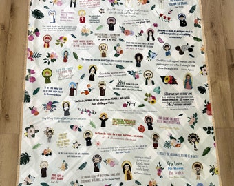 May include: A white and cream colored fleece blanket with a repeating pattern of floral designs and images of saints. The blanket features quotes from various saints, including Saint Anthony of Padua, Saint Therese of Lisieux, and Saint Agnes.