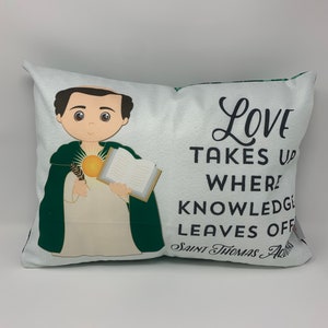 May include: A white pillow with a green and white design. The pillow features a cartoon illustration of a man holding a book and a quill. The text on the pillow reads "Love takes up where knowledge leaves off. Saint Thomas Aquinas."
