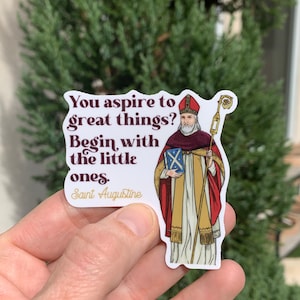 May include: A sticker featuring a cartoon illustration of Saint Augustine holding a book and a staff. The sticker has a white background with the quote "You aspire to great things? Begin with the little ones. Saint Augustine" in brown text.