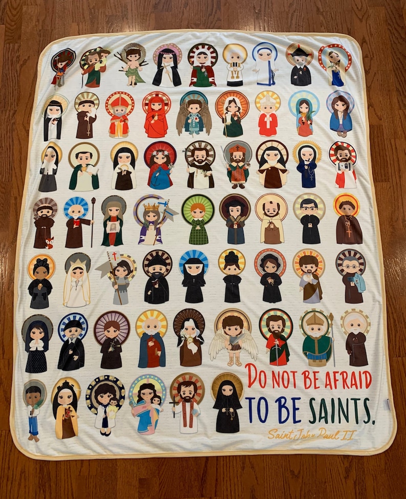 May include: A white fleece blanket with a colorful illustration of various saints. The blanket features a quote from Saint John Paul II: "Do not be afraid to be saints."