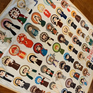 May include: A white fleece blanket with a colorful pattern of various saints. The blanket features a quote: "Do not be afraid to be saints."
