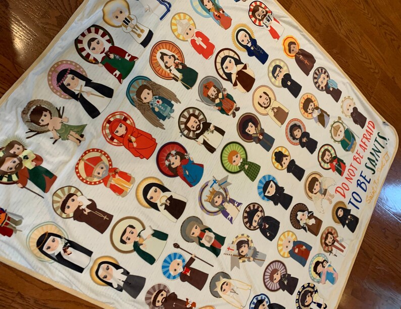 May include: A white fleece blanket with a colorful pattern of various saints. The blanket features a quote: "Do not be afraid to be saints."