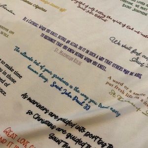 May include: A white fabric with colorful handwritten quotes about love, faith, and life. The quotes include: "The Lord has loved me, love everyone, we must, Saint Josephine", "Do not be unwise, go straight on, forgetful of self, letting the spirit of God act instead, Saint Jolie Billiart", "Be a Catholic: When you kneel before an altar, do it in such a way that others may be able to recognize that you know before whom you kneel. St. Maximilian Kolbe", "The ultimate test of your greatness is the way you treat every human being. Saint John Paul II", "As mariners are guided into port by the shining of stars, so Christians are guided to heaven by the shining of God's love. Saint Thomas Aquinas", "Great love can change the world, and it is only love that can save it."