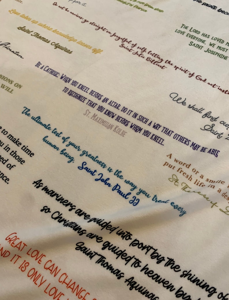 May include: A white fabric with colorful handwritten quotes about love, faith, and life. The quotes include: "The Lord has loved me, love everyone, we must, Saint Josephine", "Do not be unwise, go straight on, forgetful of self, letting the spirit of God act instead, Saint Jolie Billiart", "Be a Catholic: When you kneel before an altar, do it in such a way that others may be able to recognize that you know before whom you kneel. St. Maximilian Kolbe", "The ultimate test of your greatness is the way you treat every human being. Saint John Paul II", "As mariners are guided into port by the shining of stars, so Christians are guided to heaven by the shining of God's love. Saint Thomas Aquinas", "Great love can change the world, and it is only love that can save it."