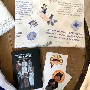 Pu&ograve; includere: Una collezione di citazioni ispiratrici sulla fede e la speranza, stampate su cartoline e un quaderno. Il quaderno ha una copertina nera con un'illustrazione colorata di santi. Le cartoline presentano illustrazioni di santi e citazioni da loro. Le citazioni includono "Amiamo la Croce e ricordiamo che non siamo soli nel portarla. Dio ci aiuta. E in Dio che ci consola, come dice San Paolo, possiamo fare qualsiasi cosa." e "Non abbandonatevi alla disperazione. Siamo il popolo di Pasqua e Alleluia &egrave; il nostro canto."
