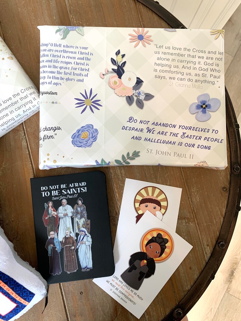 Pu&ograve; includere: Una collezione di citazioni ispiratrici sulla fede e la speranza, stampate su cartoline e un quaderno. Il quaderno ha una copertina nera con un'illustrazione colorata di santi. Le cartoline presentano illustrazioni di santi e citazioni da loro. Le citazioni includono "Amiamo la Croce e ricordiamo che non siamo soli nel portarla. Dio ci aiuta. E in Dio che ci consola, come dice San Paolo, possiamo fare qualsiasi cosa." e "Non abbandonatevi alla disperazione. Siamo il popolo di Pasqua e Alleluia &egrave; il nostro canto."