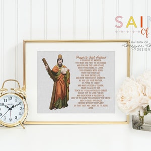 May include: A framed print featuring a religious illustration of Saint Andrew holding a cross and a book, with a prayer printed on the canvas. A gold alarm clock and a vase of white flowers are also visible.