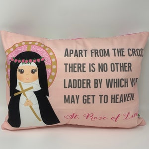 May include: A pink pillow with a quote from Saint Rose of Lima: "Apart from the cross there is no other ladder by which we may get to heaven." The pillow features an illustration of Saint Rose of Lima holding a cross.