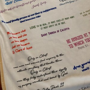 May include: A white fabric tapestry with inspirational quotes from various saints and religious figures. The quotes are written in black, red, and blue ink. The tapestry features quotes from Saint Cecilia, Saint Lucy, Saint Maximikan Kolbe, Saint Theresa of Calcutta, Saint Clare of Assisi, Pope St. John Paul II, and Pope St.
