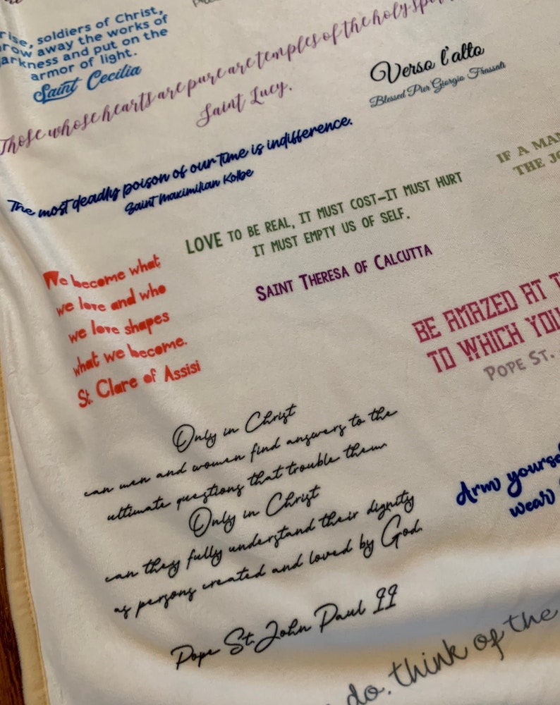 May include: A white fabric tapestry with inspirational quotes from various saints and religious figures. The quotes are written in black, red, and blue ink. The tapestry features quotes from Saint Cecilia, Saint Lucy, Saint Maximikan Kolbe, Saint Theresa of Calcutta, Saint Clare of Assisi, Pope St. John Paul II, and Pope St.