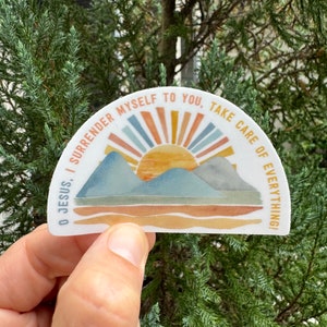May include: A white sticker with a watercolor illustration of a sunrise over mountains. The text "O Jesus, I surrender myself to you, take care of everything!" is written around the edge of the sticker.