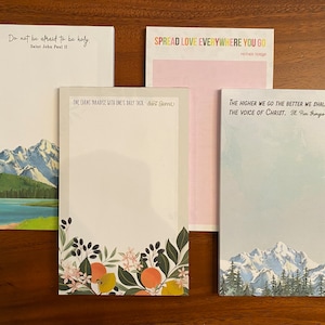 May include: Four different notepads with inspirational quotes and mountain scenery. The quotes are: "Do not be afraid to be holy. Saint John Paul II", "Spread love everywhere you go. Mother Teresa", "One earns paradise with one's daily task. Saint Gamu", and "The higher we go the better we shall hear the voice of Christ. Bl. Pier Giorgio Frassati".