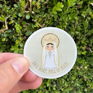 May include: A round sticker featuring an illustration of a woman in a white veil and dress, with a gold halo and crown. The text "HAIL MARY, FULL OF GRACE" is printed around the edge of the sticker. The sticker is held in a hand.