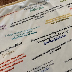 May include: A white fabric tapestry with colorful inspirational quotes from various saints and religious figures. The quotes are printed in different colors and fonts. The tapestry is a great way to add a touch of faith and inspiration to your home decor.