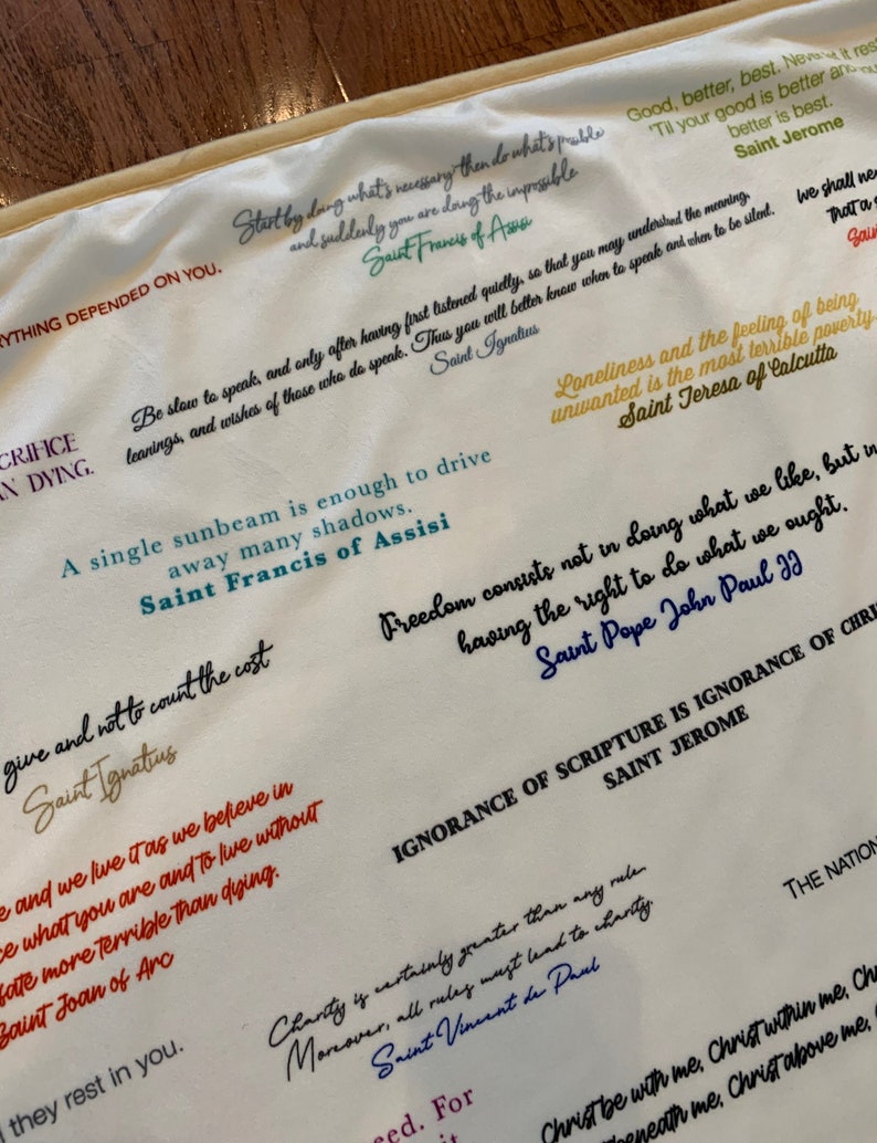 May include: A white fabric tapestry with colorful inspirational quotes from various saints and religious figures. The quotes are printed in different colors and fonts. The tapestry is a great way to add a touch of faith and inspiration to your home decor.