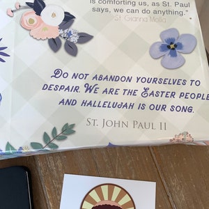 Pu&ograve; includere: Un libro con un designo floreale sulla copertina. Il testo sulla copertina recita "Non abbandonatevi alla disperazione. Siamo il popolo di Pasqua e Alleluia &egrave; il nostro canto. San Giovanni Paolo II".