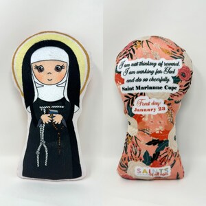 May include: A plush toy of Saint Marianne Cope, a Catholic saint, with a floral pattern on the back. The back of the toy includes a quote from Saint Marianne Cope: "I am not thinking of reward. I am working for God and do so cheerfully." The back also includes the saint's feast day: January 23.