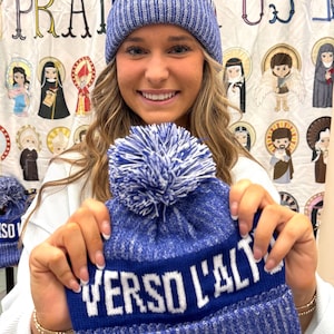 May include: A blue and white knit beanie with a white pom pom and the text "VERSO L'ALTO" in white letters. The beanie is being held by a person.