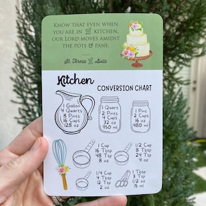 May include: A green and white kitchen conversion chart with illustrations of measuring cups, spoons, and a whisk. The chart includes measurements in cups, tablespoons, teaspoons, ounces, and milliliters. The quote "Know that even when you are in the kitchen, our Lord moves amidst the pots & pans. St. Teresa of Avila" is printed at the top.