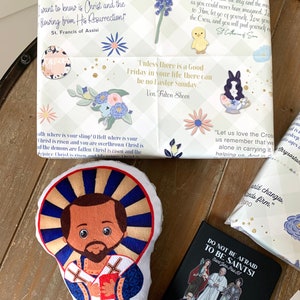 Pu&ograve; includere: Un giocattolo di peluche di San Francesco d'Assisi, un santo cattolico, con una tunica blu e bianca e un'aureola. Il giocattolo &egrave; su una superficie di legno con altri oggetti religiosi, tra cui un regalo avvolto con una citazione di Santa Caterina da Siena, un piccolo libro con il testo "Non abbiate paura di essere santi!" e un regalo avvolto con una citazione di San Francesco d'Assisi.