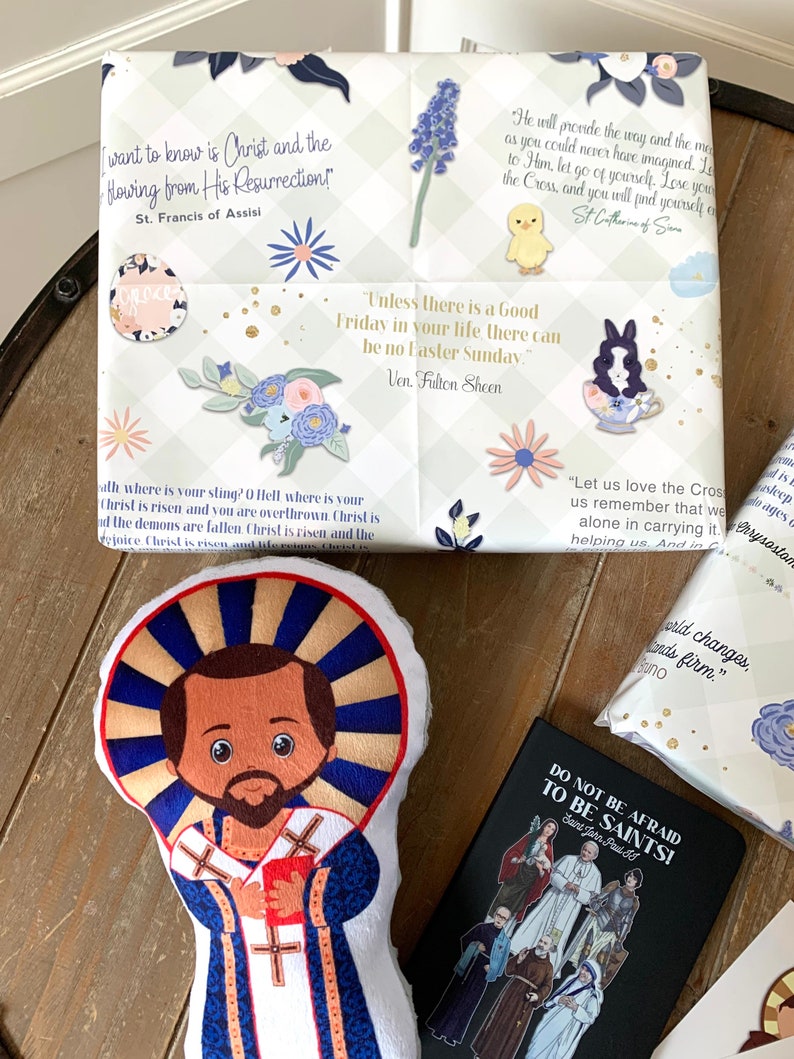 Pu&ograve; includere: Un giocattolo di peluche di San Francesco d'Assisi, un santo cattolico, con una tunica blu e bianca e un'aureola. Il giocattolo &egrave; su una superficie di legno con altri oggetti religiosi, tra cui un regalo avvolto con una citazione di Santa Caterina da Siena, un piccolo libro con il testo "Non abbiate paura di essere santi!" e un regalo avvolto con una citazione di San Francesco d'Assisi.