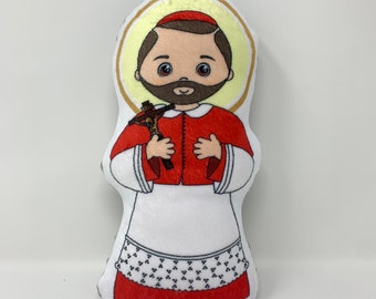Könnte beinhalten: Ein Plüschpuppe, die eine religiöse Figur darstellt, mit rotem Gewand, weißer Unterwäsche und einem Heiligenschein. Die Puppe hält ein kleines Kruzifix. Das Design umfasst einen braunen Bart und Haare mit einem lächelnden Ausdruck. Die Puppe ist etwa 20 cm groß.