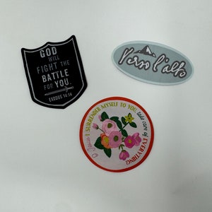 May include: Three iron-on patches with inspirational messages. The first patch is black with white text that reads "God will fight the battle for you. Exodus 14:14". The second patch is light blue with white text that reads "Ven l'alto". The third patch is orange with a floral design and white text that reads "To Jesus I surrender myself to you, take care of everything!"