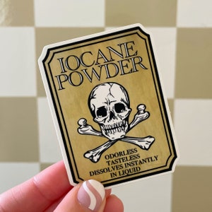 May include: A sticker with a vintage design featuring the text "IOCANE POWDER" above a skull and crossbones. The sticker also includes the text "ODORLESS TASTELESS DISSOLVES INSTANTLY IN LIQUID" on a gold background with a black border.