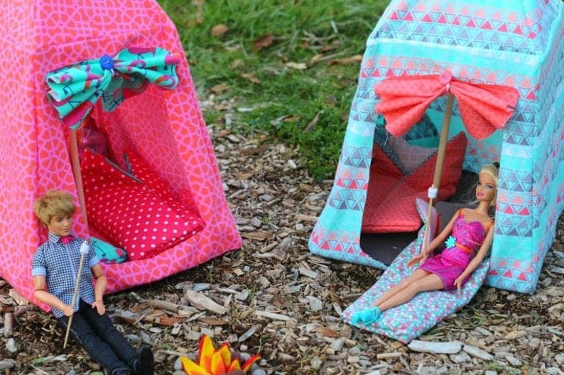 May include: Two colourful fabric tents for dolls, one pink with white polka dots and one blue with a geometric pattern. A doll is inside the blue tent, and another doll is outside the pink tent holding a stick.