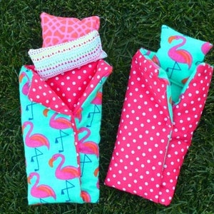May include: Two pink and green sleeping bags with flamingo patterns and white polka dots. One sleeping bag has a pink pillow with a flamingo design and a white crocheted pillow on top. Both sleeping bags are on a green grassy surface.
