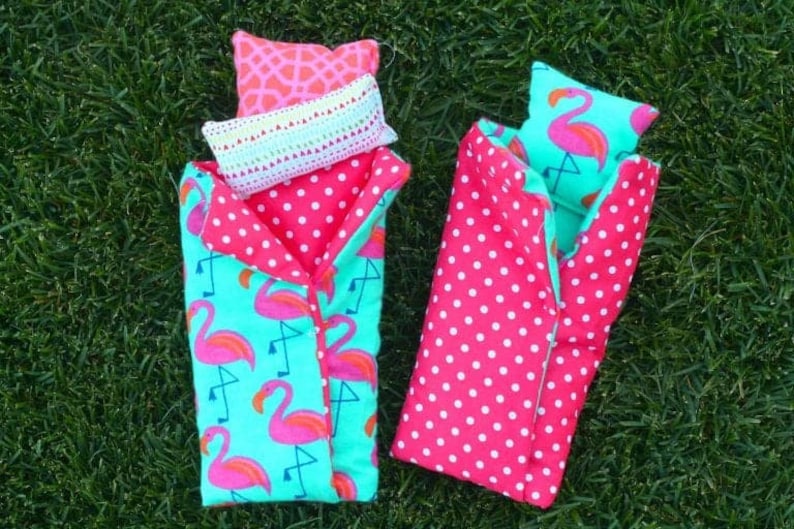 May include: Two pink and green sleeping bags with flamingo patterns and white polka dots. One sleeping bag has a pink pillow with a flamingo design and a white crocheted pillow on top. Both sleeping bags are on a green grassy surface.