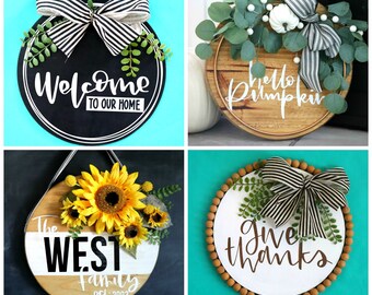 May include: Four different round wooden signs with various decorations and text. The first sign is black with white text that says "Welcome to our home" and has a black and white striped bow and greenery. The second sign is natural wood with white text that says "Hello Pumpkin" and has a white bow, greenery, and white berries. The third sign is a split wood sign with white text that says "The West Family est. 2003" and has sunflowers and greenery. The fourth sign is white with brown text that says "Give Thanks" and has a black and white striped bow, greenery, and brown beads around the edge.