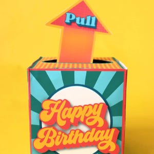 May include: A colorful birthday gift box with a pull tab arrow. The box is teal with a white circle and the words "Happy Birthday" in orange and yellow. The arrow is orange with the word "Pull" in blue.