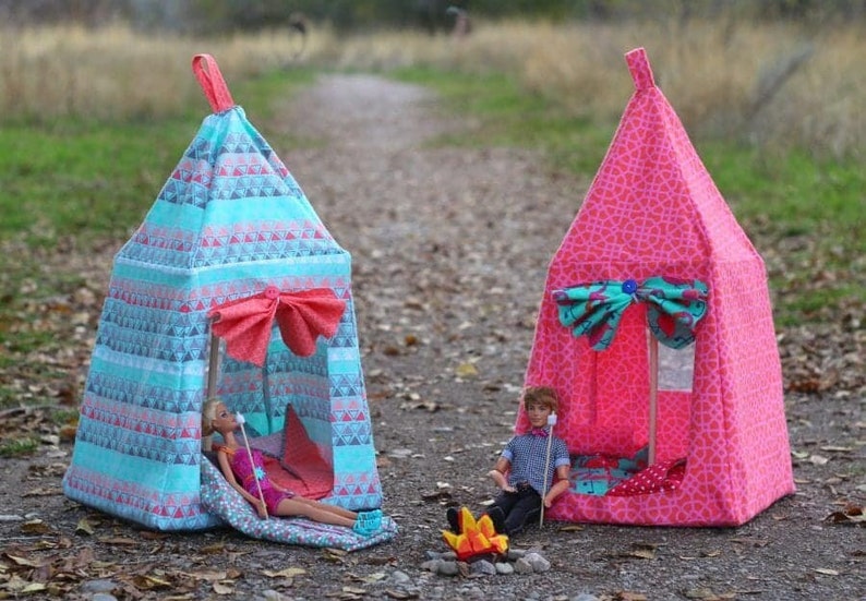 May include: Two colourful fabric teepees with dolls inside. The teepees are decorated with geometric patterns and have a bow at the entrance. One teepee is blue and pink, the other is pink and teal. The dolls are sitting by a small campfire.