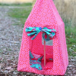 May include: A pink and turquoise play tent with a geometric pattern. The tent has a flap that can be tied closed with a bow. The tent is decorated with a flamingo print. The tent is sitting on a bed of wood chips.