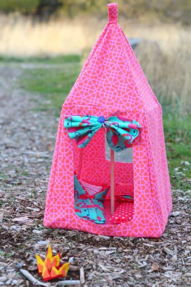 May include: A pink and turquoise play tent with a geometric pattern. The tent has a flap that can be tied closed with a bow. The tent is decorated with a flamingo print. The tent is sitting on a bed of wood chips.