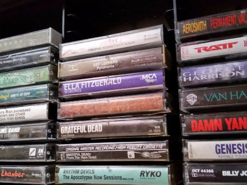 May include: A close-up of a shelf with a variety of music cassette tapes. The tapes are arranged vertically and feature different artists and album titles, including "Def Leppard", "Ella Fitzgerald", "Van Halen", "Ratt", "George Harrison", "Genesis", "Damn Yankees", "Billy Joel", "Grateful Dead", and "Rhythm Devils".
