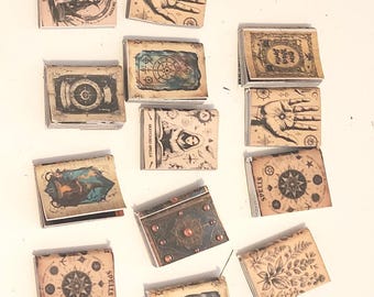 May include: A collection of small, vintage-style matchboxes with various illustrations and text. The designs include celestial maps, hands, botanical prints, and portraits, all in a muted color palette. Some boxes have the word "SPELLS" printed on them.