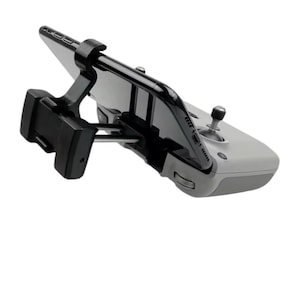 May include: A grey drone controller with a black phone holder attached. The phone holder is in a vertical position and the phone is not visible. The controller has a joystick and buttons.