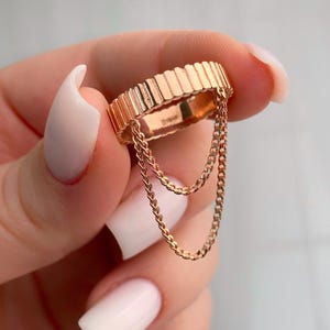 May include: A rose gold ring with a textured band and two dangling chains.