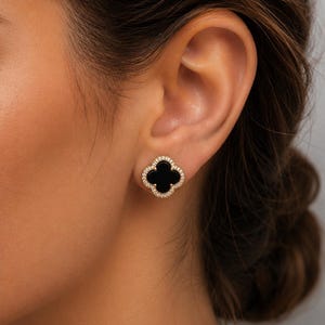 May include: A stud earring featuring a black four-leaf clover shape set in a gold-toned metal frame. The clover is surrounded by a border of small, clear, sparkling stones that follow the scalloped edges of the design, creating a contrast.