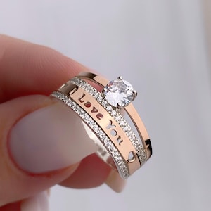 May include: A silver and rose gold ring set with a clear cubic zirconia stone. The band of the ring has the words "I Love You" engraved on it.